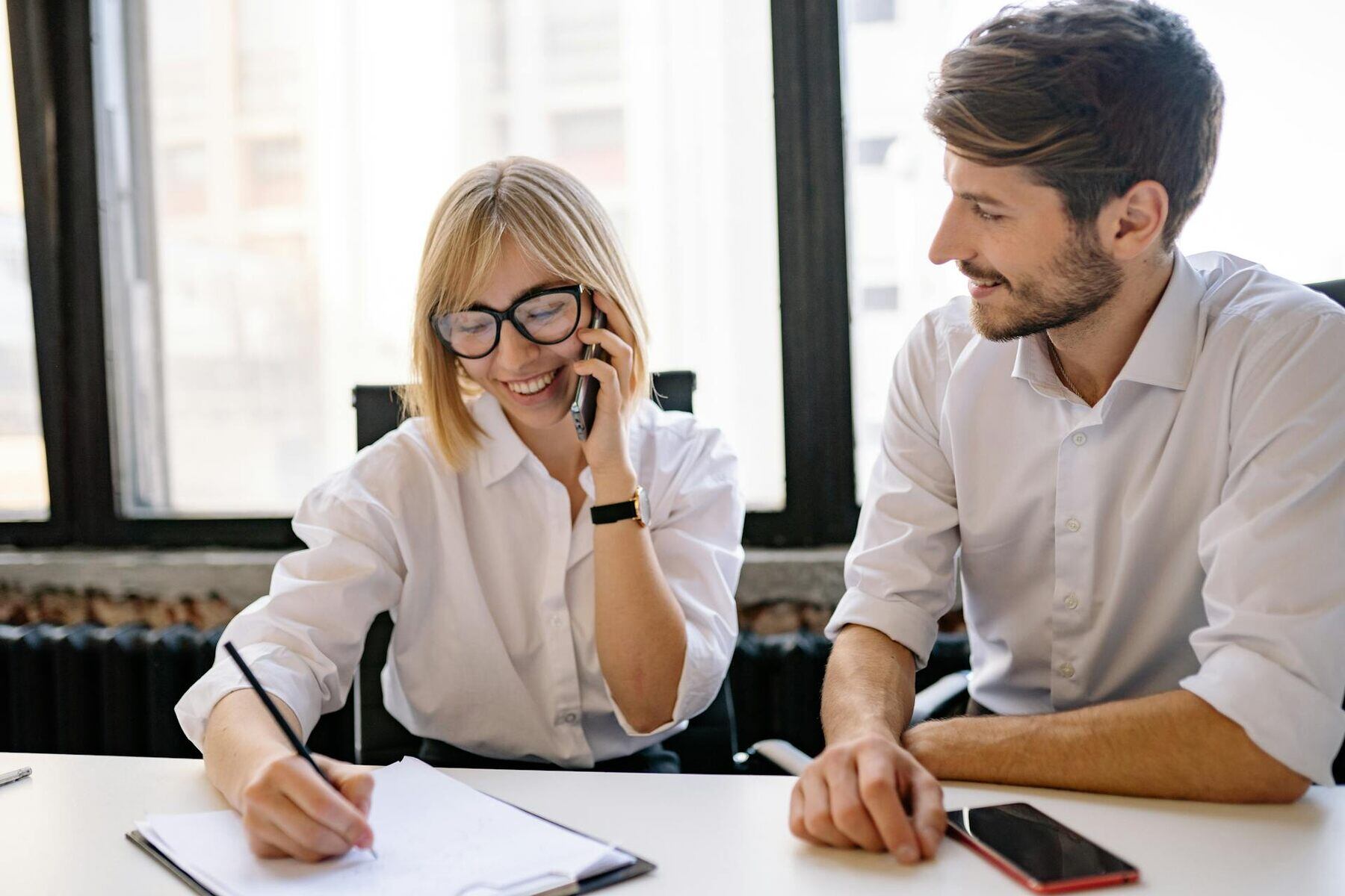 A man and woman engaged in a conversation while sitting at a desk and using a mobile phone